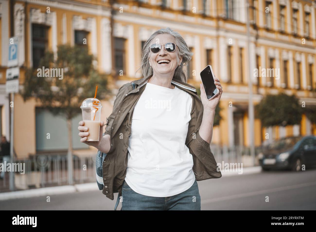 Glückliche ältere Frau, die draußen lächelt. Sie spricht am Telefon, engagiert sich in moderner Kommunikation und genießt erfrischende Getränke Stockfoto