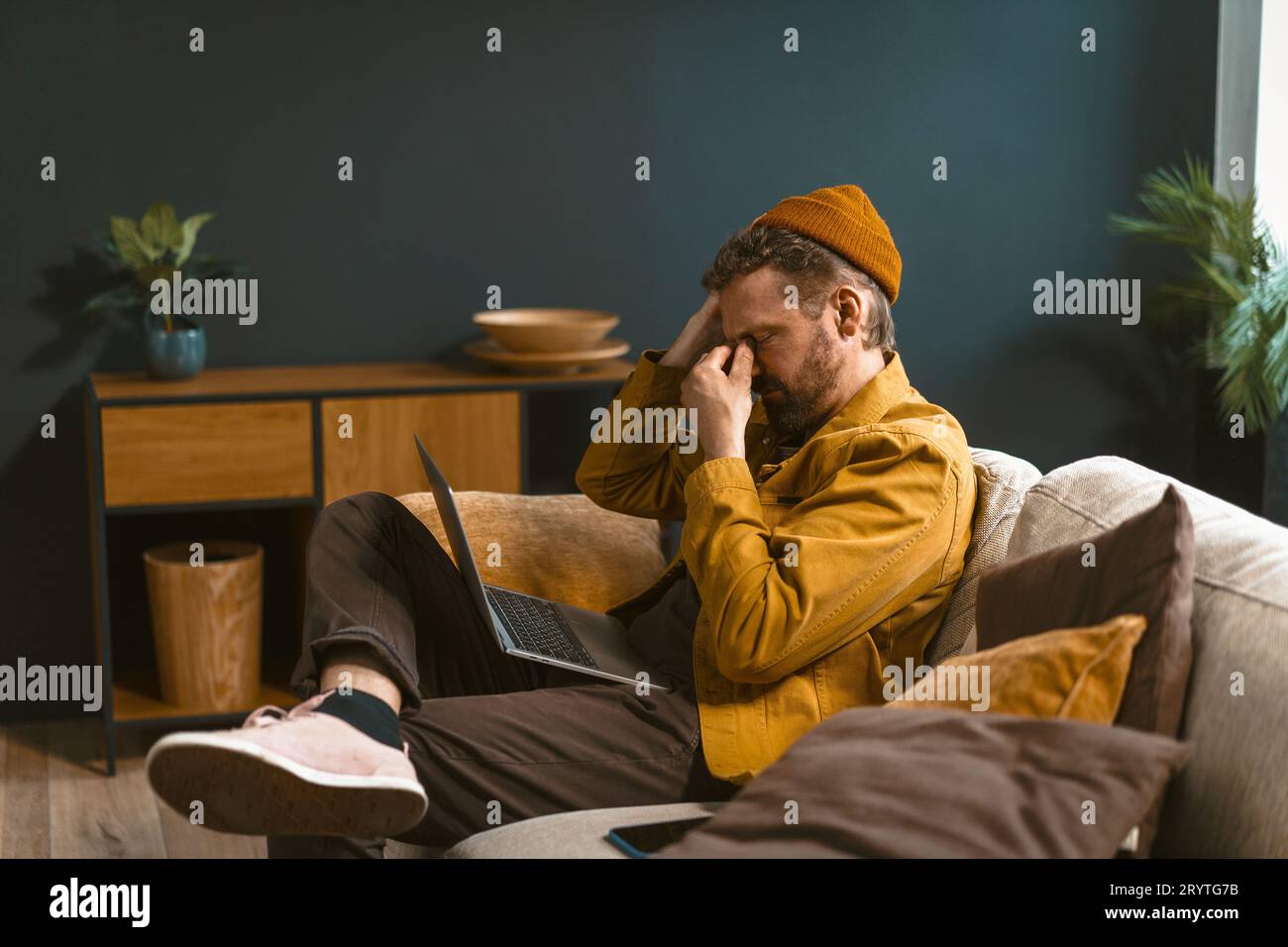 Ein trauriger und müder Mann sitzt zu Hause auf einem Sofa. Überwältigt von schlechten Nachrichten oder Herausforderungen, hält er seinen Kopf mit geschlossenem Ei Stockfoto