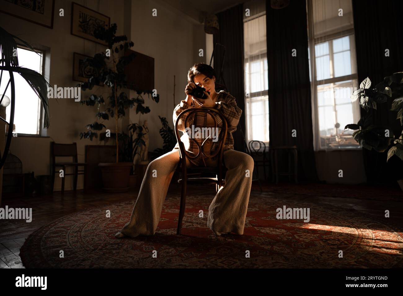 Ein junger Erwachsener sitzt in einem Raum, der von einem einzigen Fenster beleuchtet wird, mit einer Kamera in den Händen Stockfoto