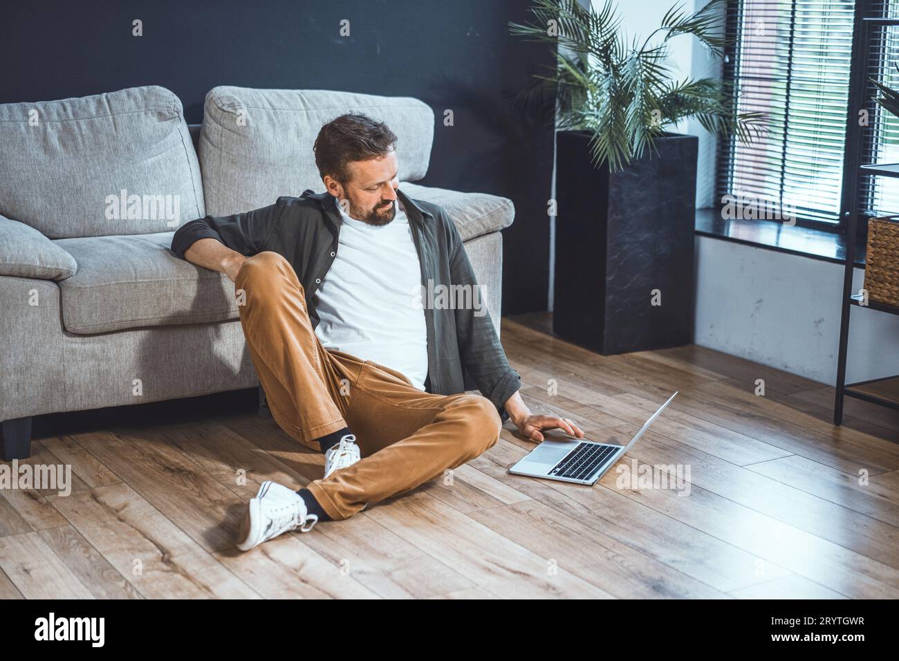 Fauler und gutaussehender Mann, der Zeit zu Hause verbringt. Mann sitzt bequem und surft im Internet auf einem Laptop. Mit entspannter Körperhaltung und lässig Stockfoto
