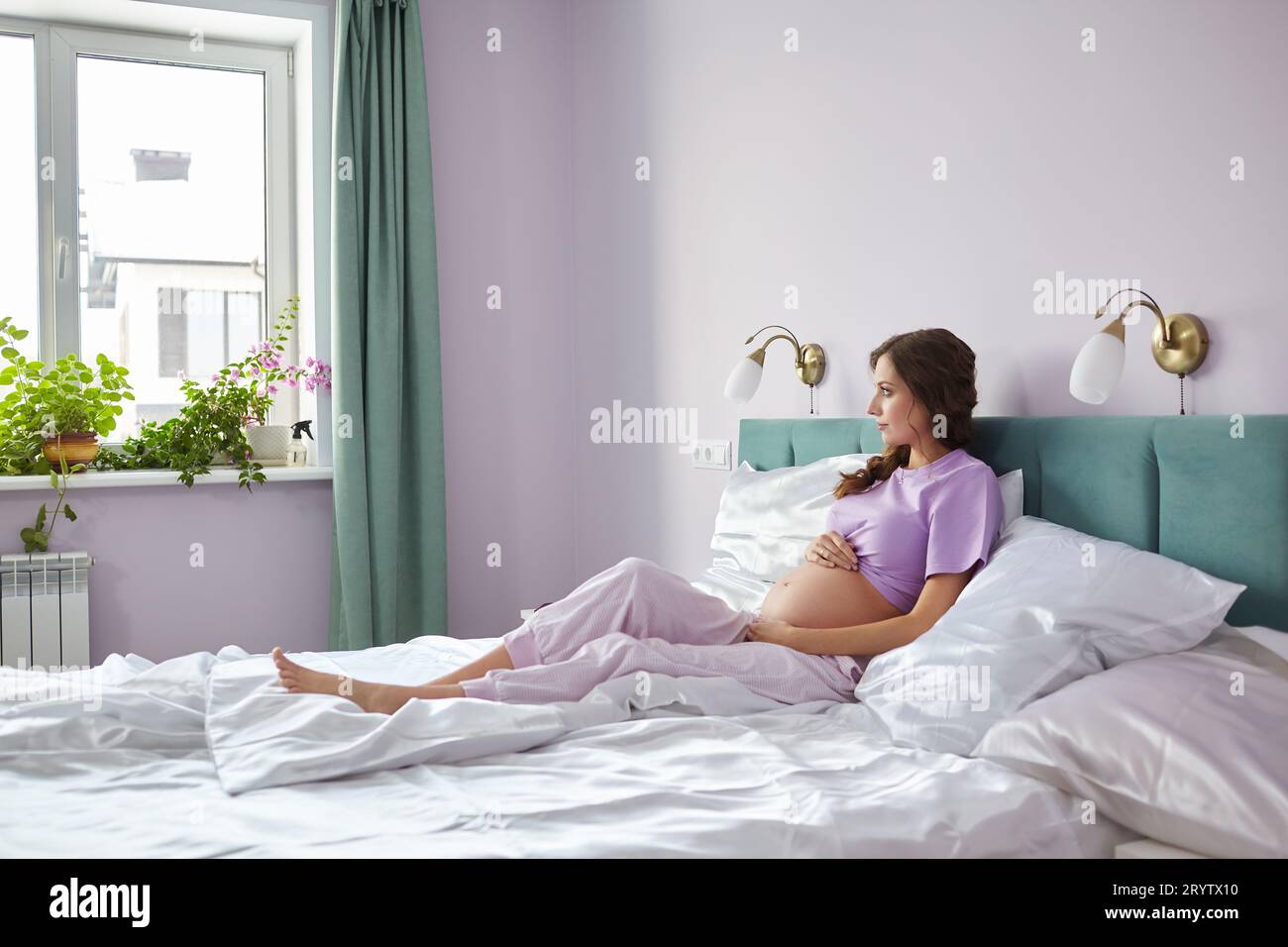 Eine schwangere Frau sitzt im Bett ihres Schlafzimmers und schaut aus dem Fenster. Frau genießt ihren Schwangerschaftszustand. Stockfoto