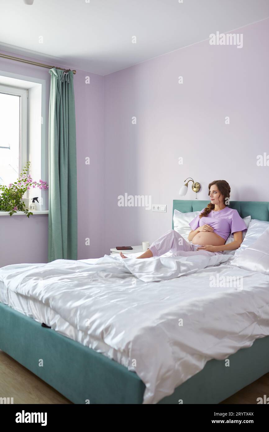 Eine schwangere Frau sitzt im Bett ihres Schlafzimmers und schaut aus dem Fenster. Frau genießt ihren Schwangerschaftszustand. Stockfoto
