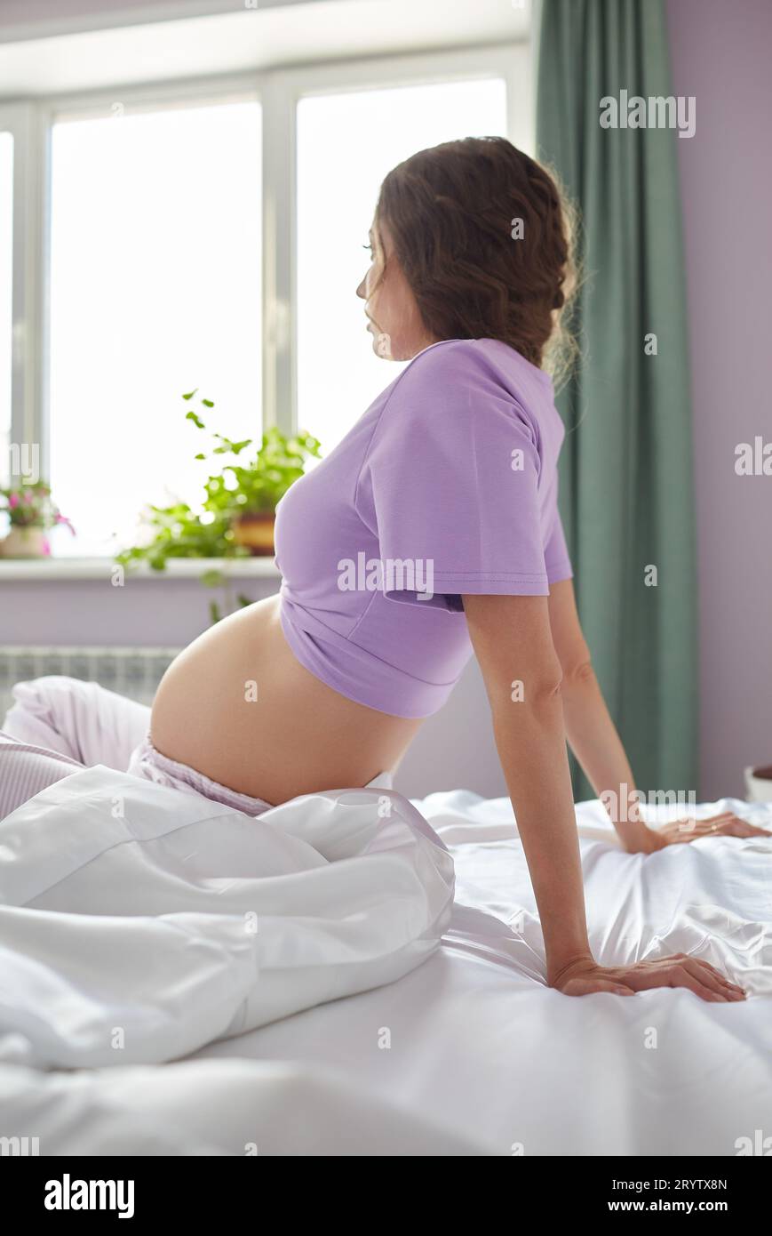 Eine schwangere Frau sitzt im Bett ihres Schlafzimmers und schaut aus dem Fenster. Frau genießt ihren Schwangerschaftszustand. Stockfoto