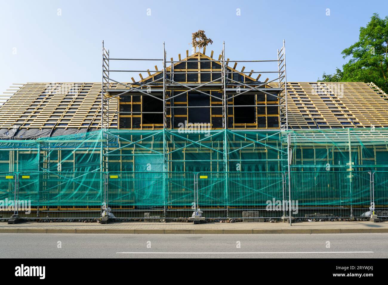 Vorderansicht der Fassade eines Holzhauses, das gerade renoviert wird, mit Gerüsten und Sicherheitsnetz, neuer Dachdrehung, temporärem Metallzaun Stockfoto