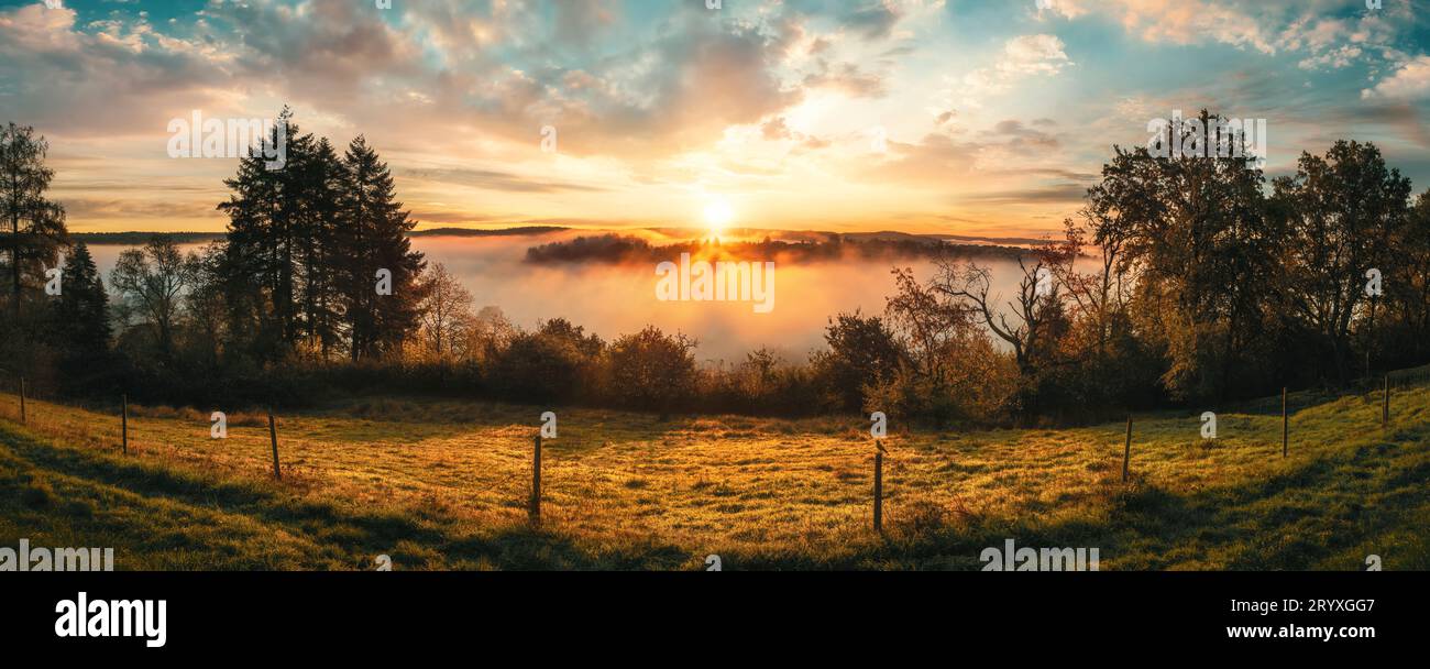Herrlicher Sonnenaufgang über ländlicher Idylle. Ein farbenfrohes Panorama mit romantischem, dramatischem Himmel, sonnendurchflutetem Nebel im Tal und Baumsilhouetten Stockfoto