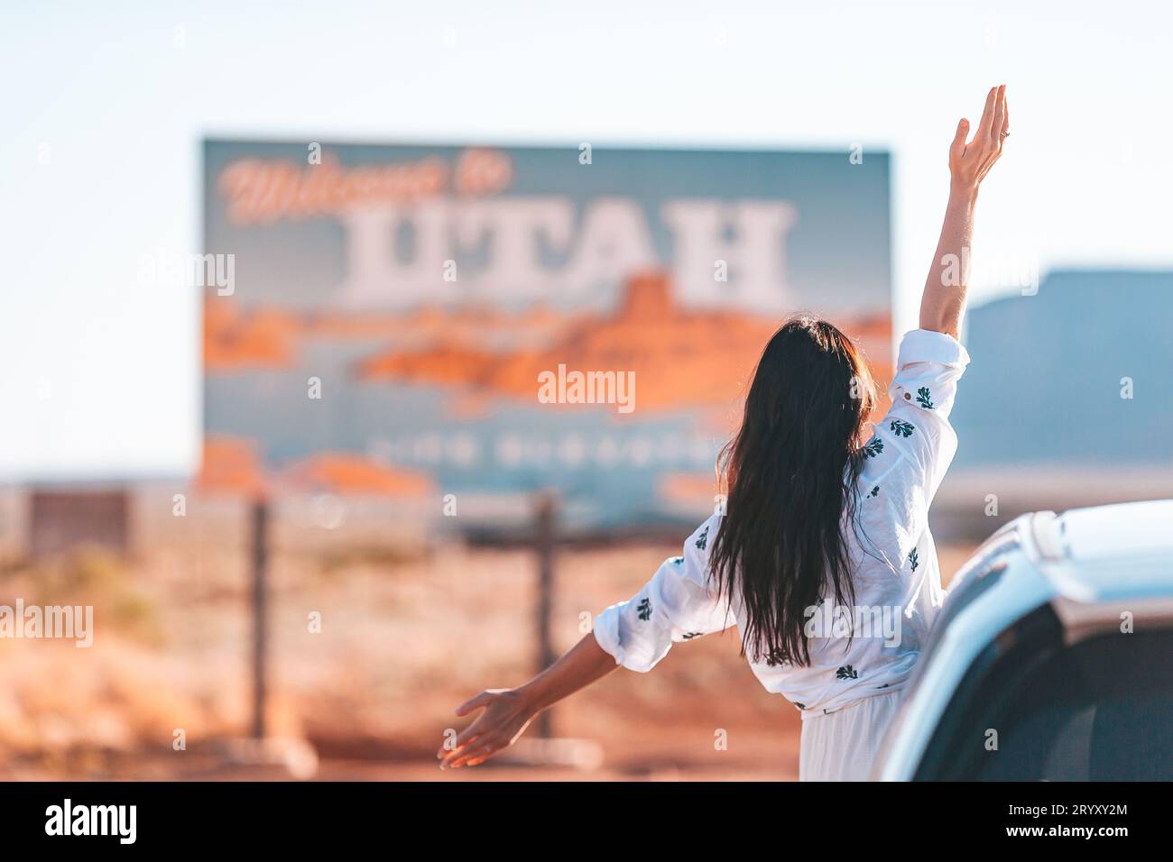 Wunderschöne Frau auf ihrer Reise am Auto vor dem Hintergrund des Hinweises Welcome to Utah State Border direkt im Monument Valley Stockfoto