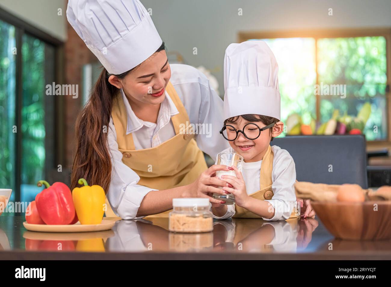 Asiatische Mutter hilft süßen kleinen Jungen, der Milch im Glas in der Küche zu Hause in Koch-Uniform trinkt. Die Menschen leben und sind glücklich Stockfoto