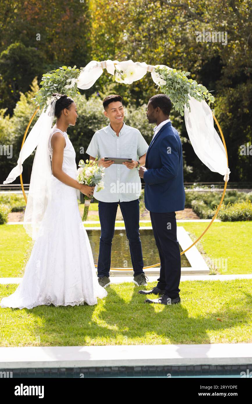 Ein vielseitiger männlicher Beamter, Braut und Bräutigam bei der Hochzeitszeremonie im sonnigen Garten Stockfoto