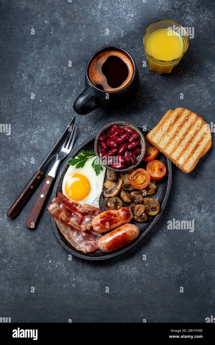 Englisches Frühstück mit Spiegeleiern, Würstchen, Speck, Pudding, Bohnen, Toast und Kaffee, dunkler Hintergrund Stockfoto