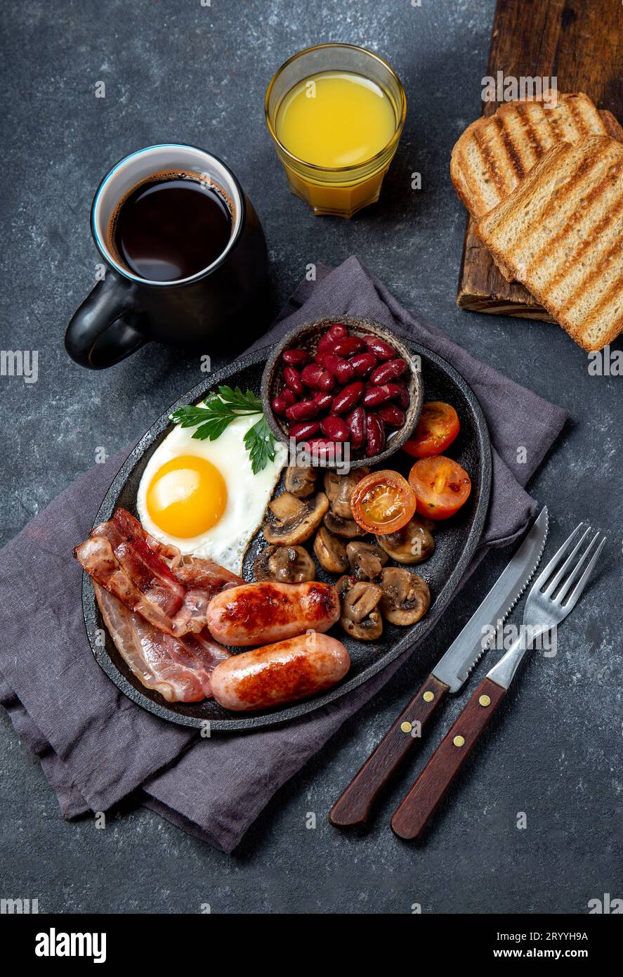 Englisches Frühstück mit Spiegeleiern, Würstchen, Speck, Pudding, Bohnen, Toast und Kaffee, dunkler Hintergrund Stockfoto