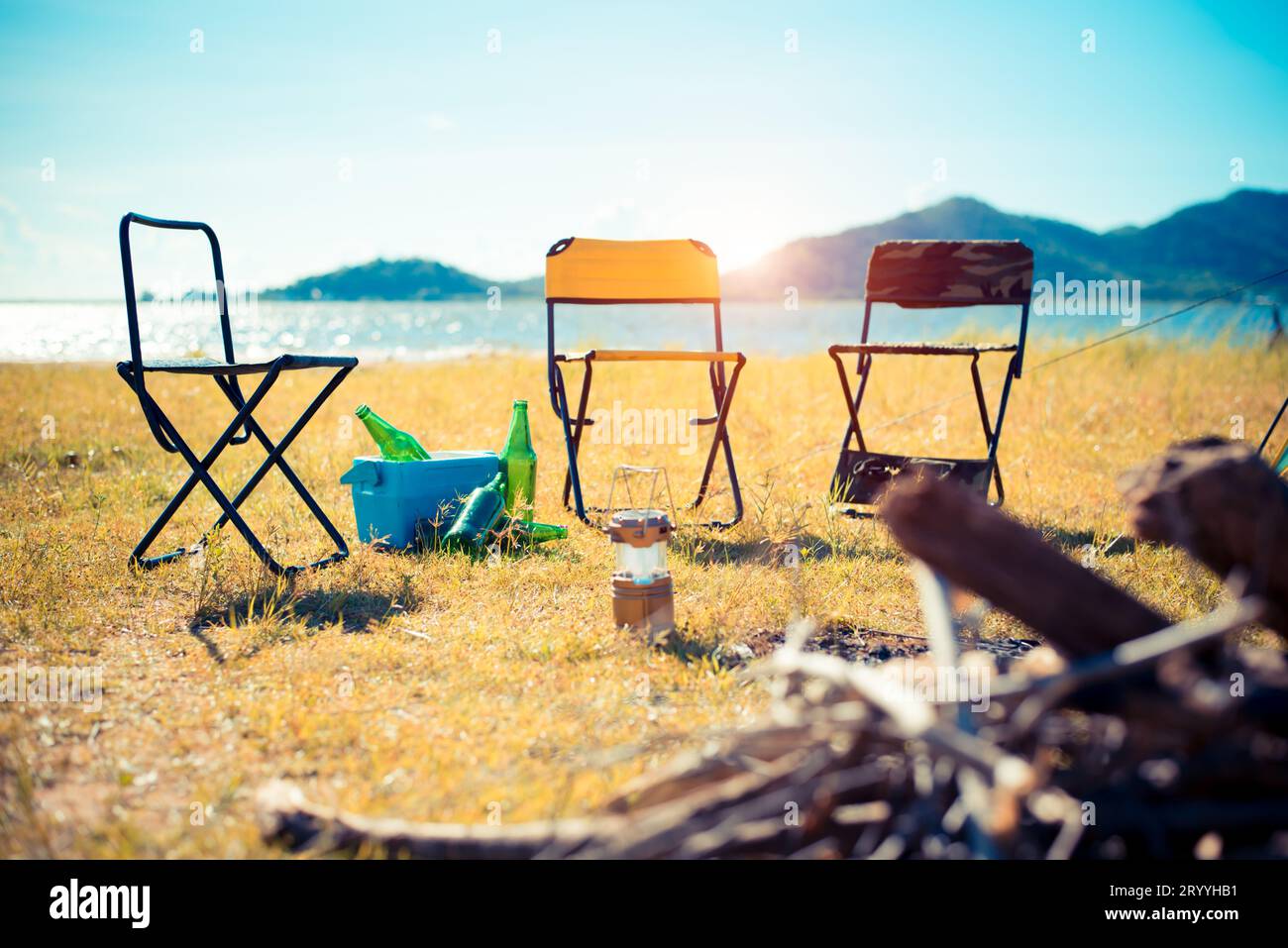 Drei Picknickstühle auf der Wiese. Lagerfeuer im Vordergrund und See mit Berg im Hintergrund. Camping- und Reisekosten Stockfoto