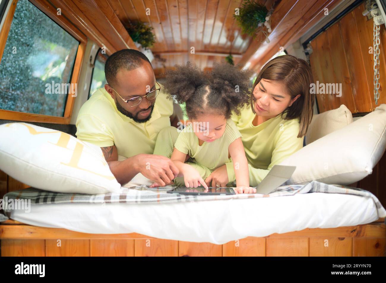 Auf der Rückseite eines weißen klassischen Minibusses spielen kleine Kinder und erfahren auf ihrem Tablet mehr über die Welt, während sich die Eltern um Hilfe kümmern. Stockfoto