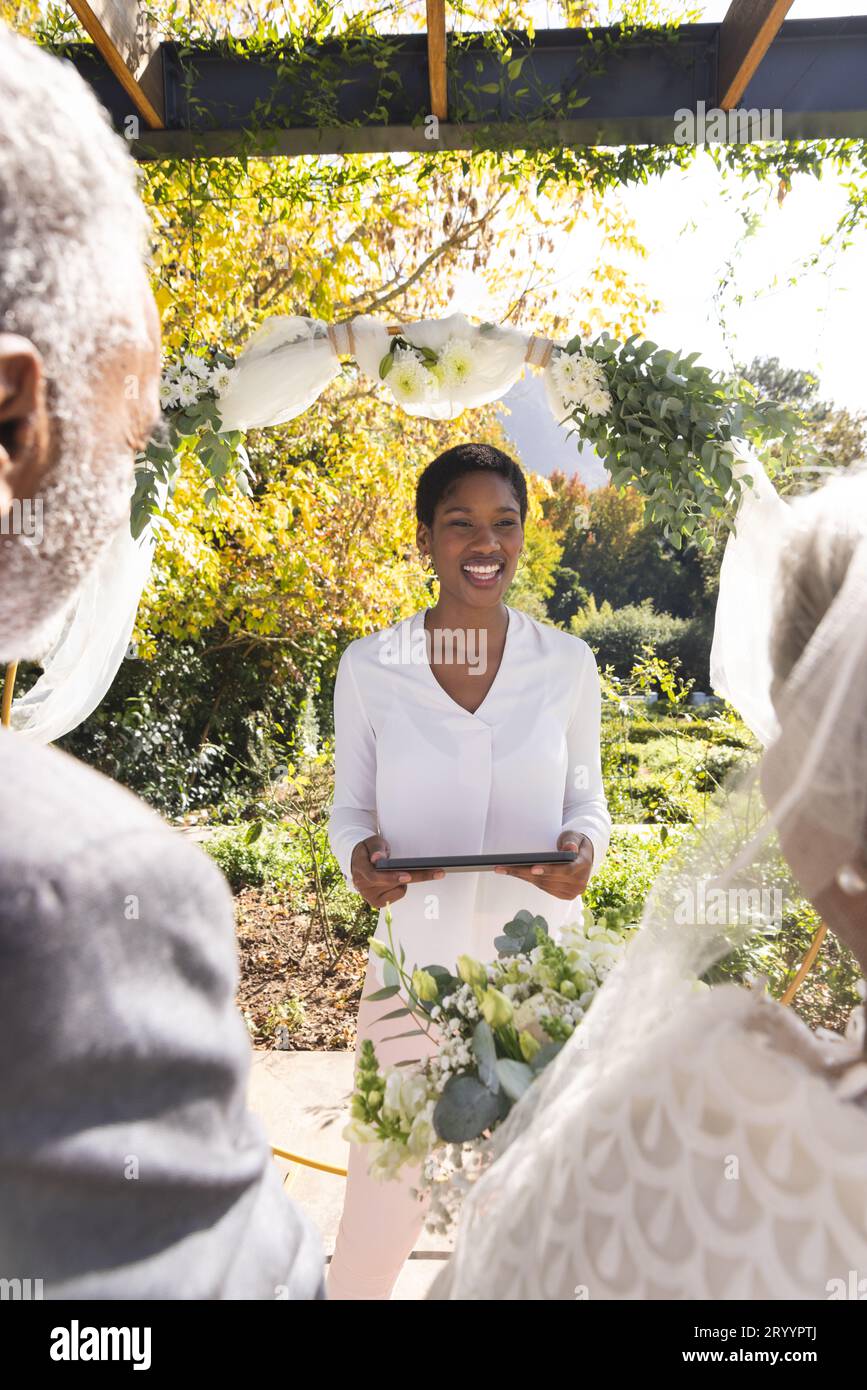 Glückliche, birassistische Frau, Ehefrau, Beamtin mit Tablet und Seniorenpaar auf der Hochzeit im sonnigen Garten Stockfoto