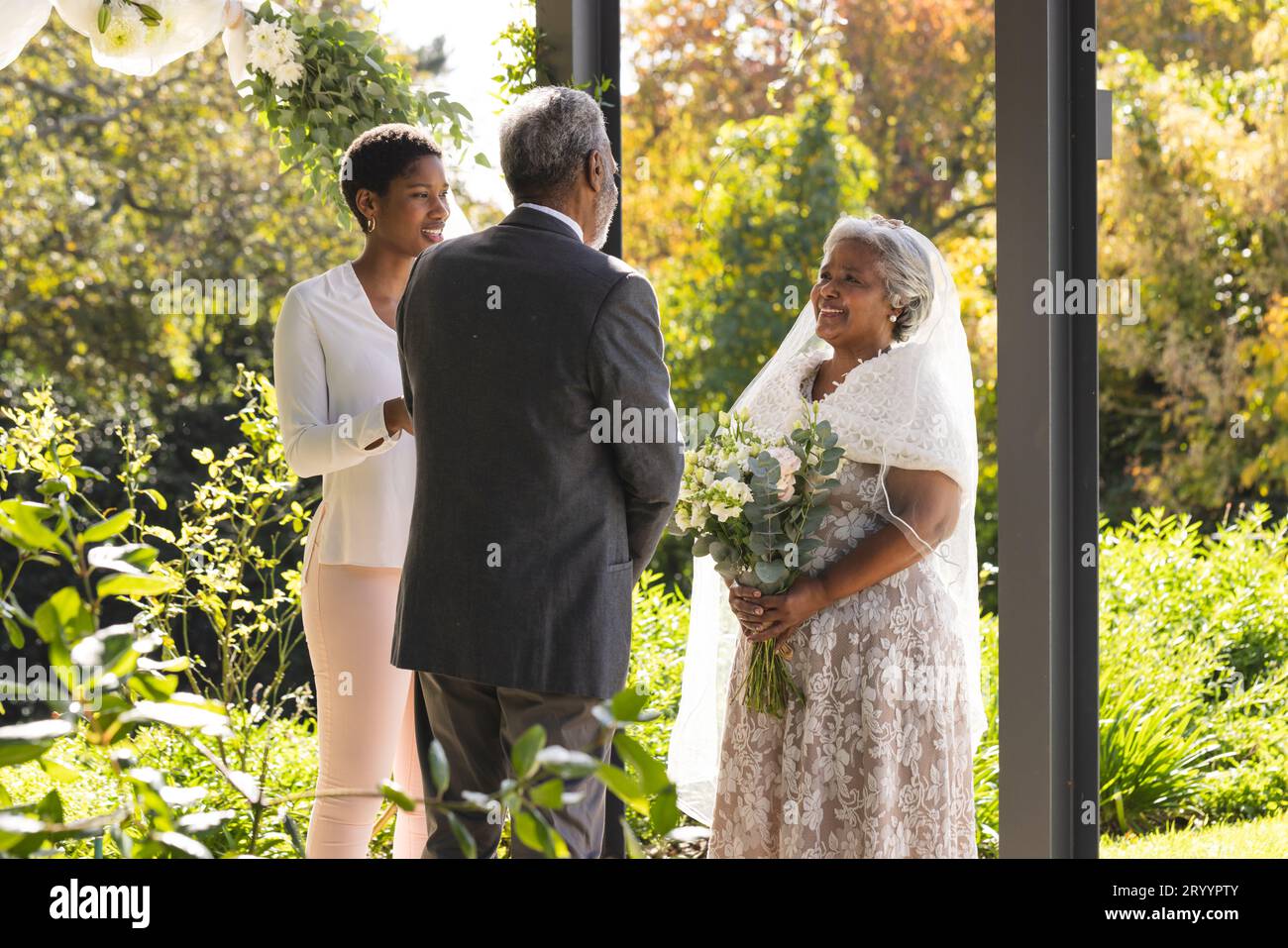 Glückliche birassische weibliche Ehestandsleiterin und Seniorenpaar während der Hochzeit im sonnigen Garten Stockfoto