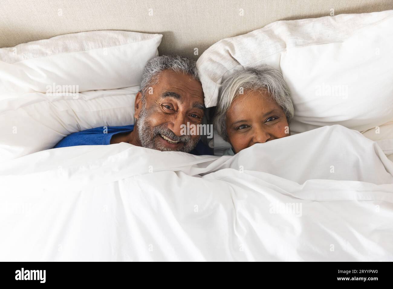 Porträt eines glücklichen älteren, birassischen Paares, das zu Hause im Bett lag und mit Bettwäsche bedeckt war Stockfoto