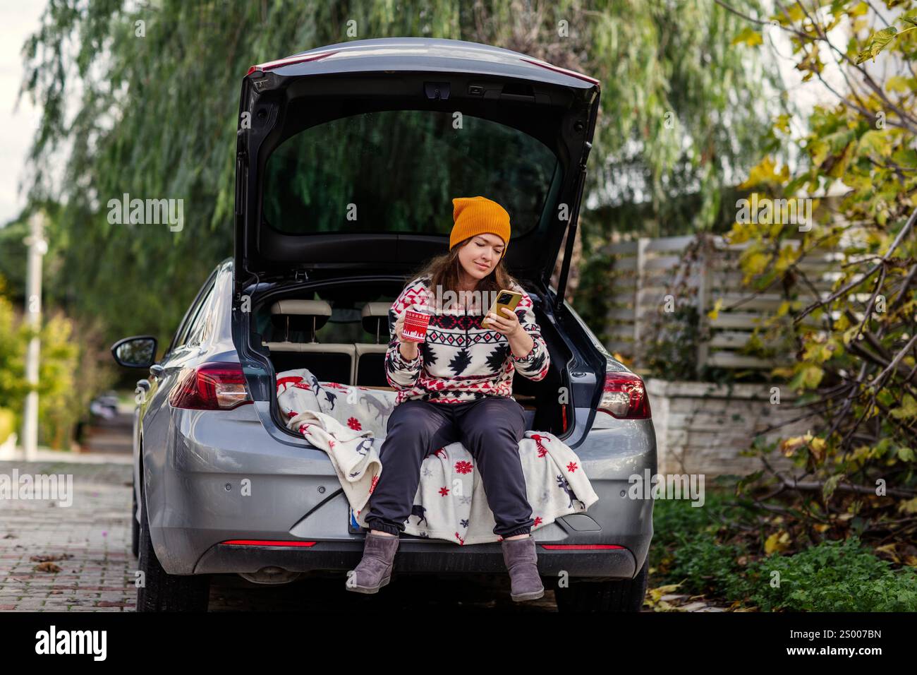 Frau sitzt mit Kaffee und Handy im Kofferraum Stockfoto