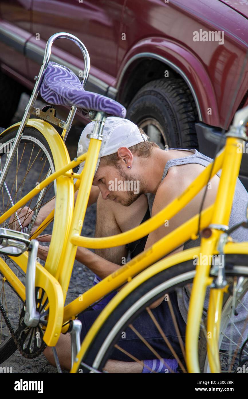 Mann repariert draußen ein gelbes Fahrrad, sitzt neben einem geparkten Auto Stockfoto