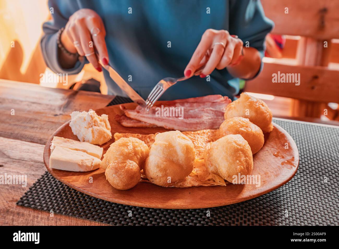 Eine Frau schneidet Speck mit Besteck, ein komplettes ländliches Frühstück mit Käse, Rührei und Priganice Donuts auf einem Holztablett in einer Berghütte Stockfoto