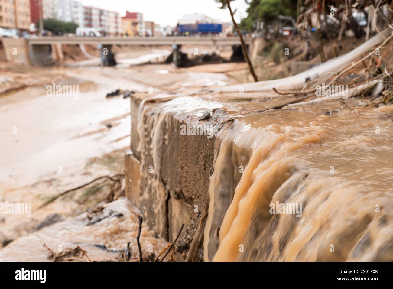 DANA-Flutungseffekte. Überschwemmungseinflüsse, Valencia, Spanien. Schlammiges Wasser, das an den Flussufern herunterfällt, mit zerstörter Stadt im Hintergrund Stockfoto