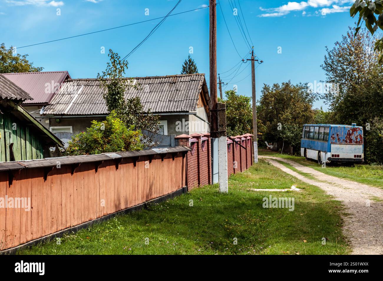 Ein blauer Bus fährt eine Feldstraße vor einem Haus entlang. Die Straße ist von Bäumen und Gras umgeben Stockfoto