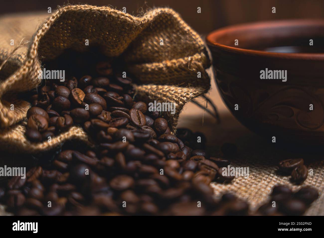 Nahaufnahme eines Sacksacks, der geröstete Kaffeebohnen neben einer braunen Keramikschale schwarzen Kaffees auf einer strukturierten Sackfläche verschüttet. Stockfoto