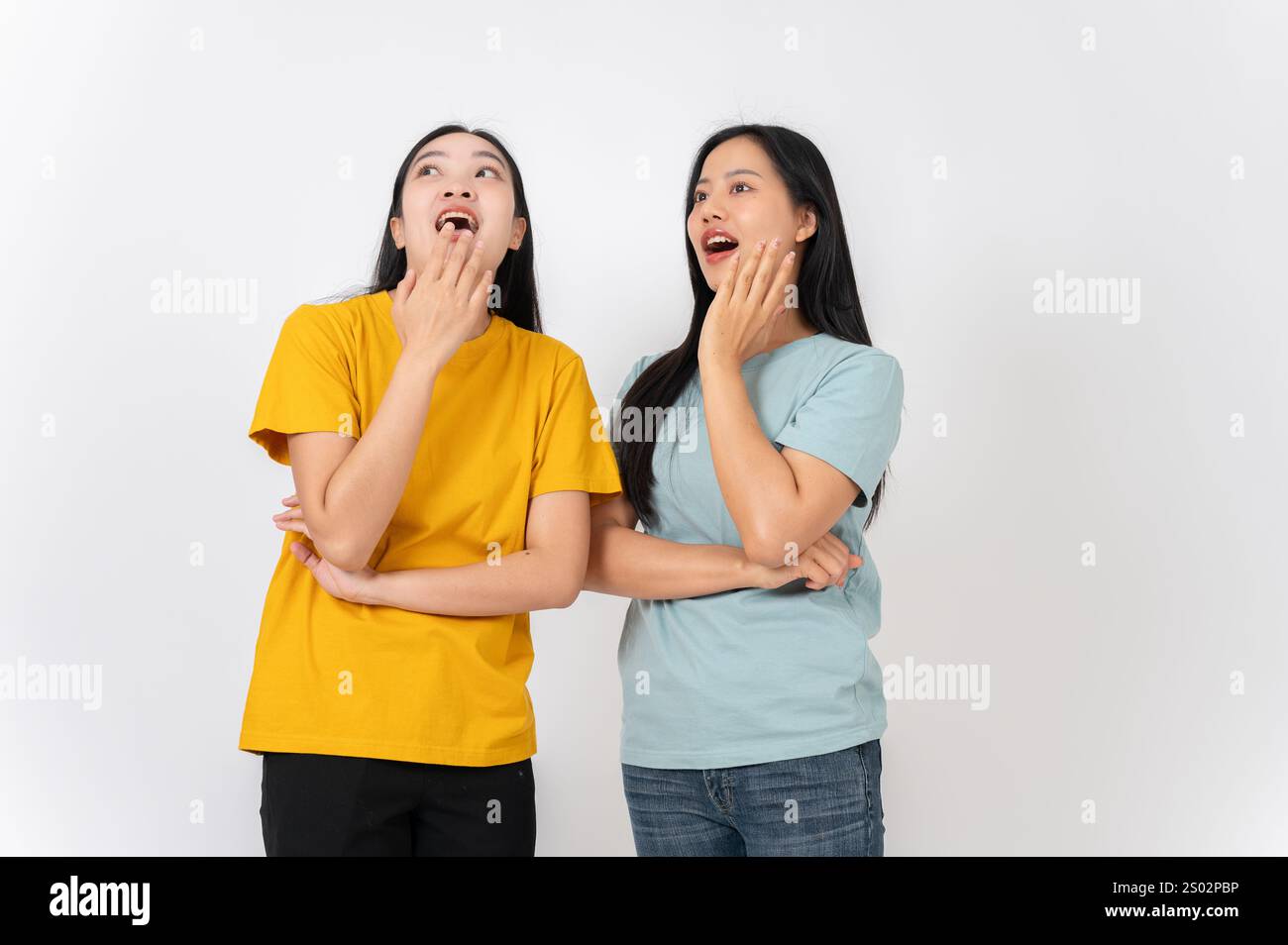 Zwei staunende und aufgeregte asiatische Frauen in lässiger Kleidung stehen vor weißem Studiohintergrund, bedecken den Mund und schauen nach oben. Überrascht B Stockfoto