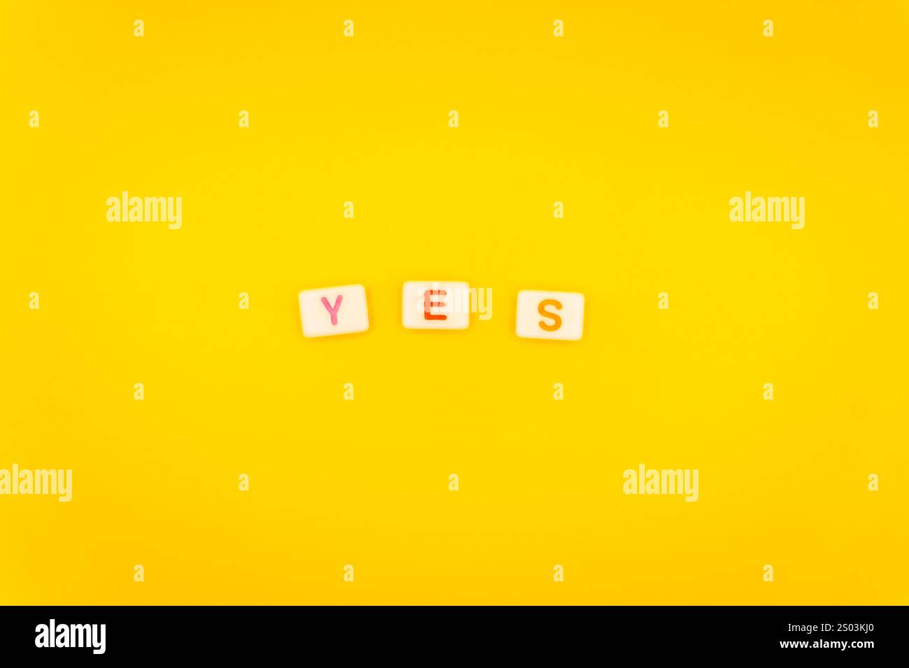Minimalistische Anordnung von Buchstabensteinen mit der Schreibweise „Ja“ mit bunten Buchstaben auf hellgelbem Hintergrund, die Affirmation und Positivität symbolisieren. Stockfoto