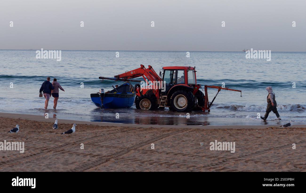 Der Traktor transportiert eine blaue Angelprahlerei vom Strand bis zum Meer im Dorf Taghazout, Marokko. Dezember 2024 Stockfoto
