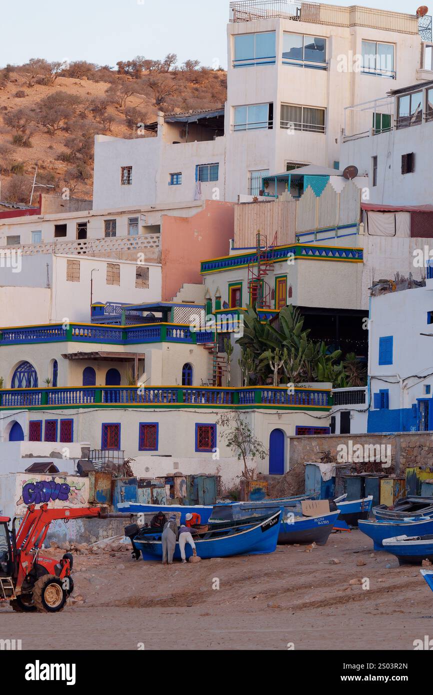 Blaue Fischerboote und ein Traktor an einem Sandstrand mit dem Dorf Taghazout dahinter, Marokko. Dezember 2024 Stockfoto