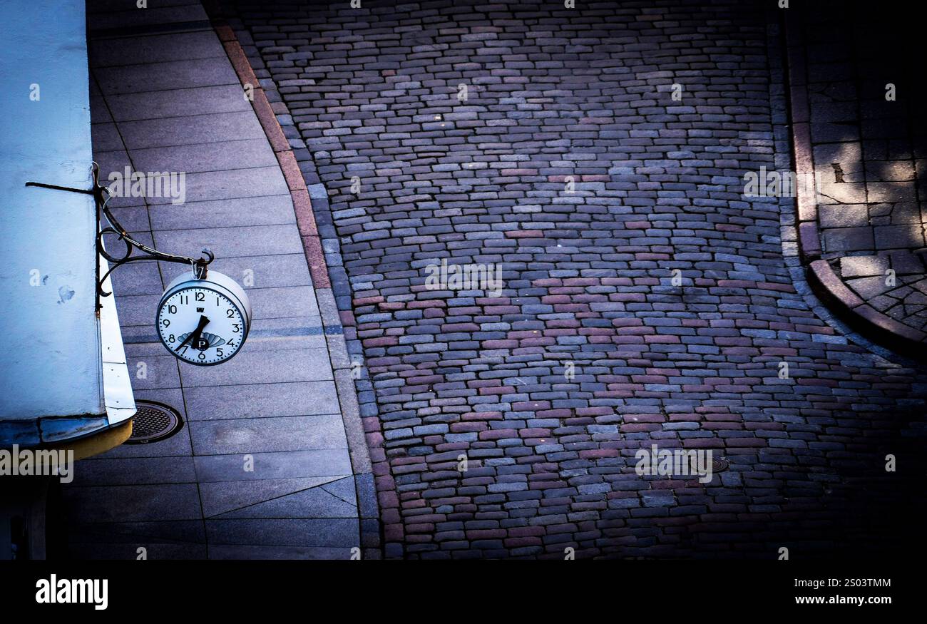 Eine Vintage-Uhr, die an einer Wand in der Nähe einer kopfsteingepflasterten Straße hängt und die komplizierten Muster der Steine zeigt. Die Szene wird mit weichem Licht beleuchtet Stockfoto