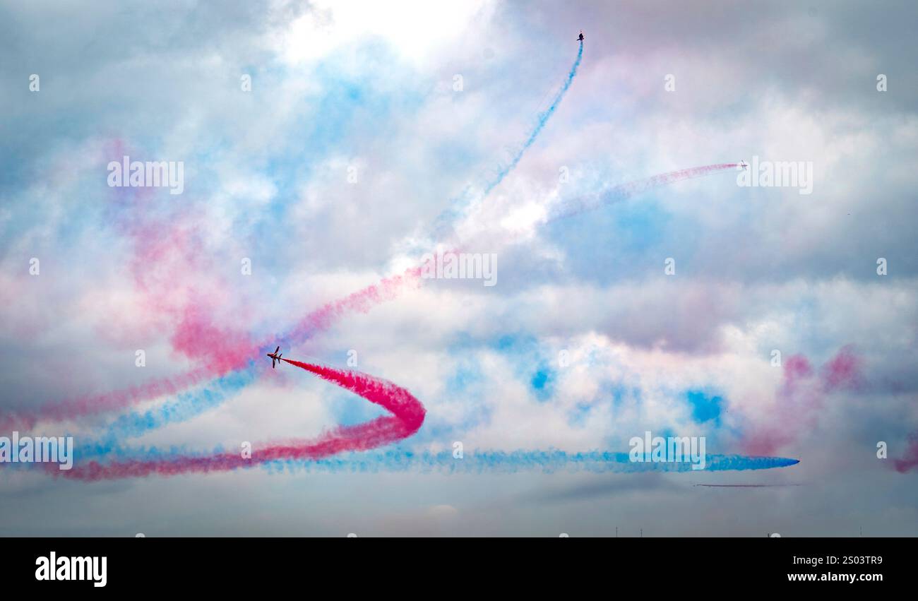 Luftbild mit Flugzeugen, die bunte Rauchpfade am Himmel erzeugen. Der Rauch ist in Rot- und Blautönen gehalten und bildet dynamische Muster gegen eine Wolke Stockfoto