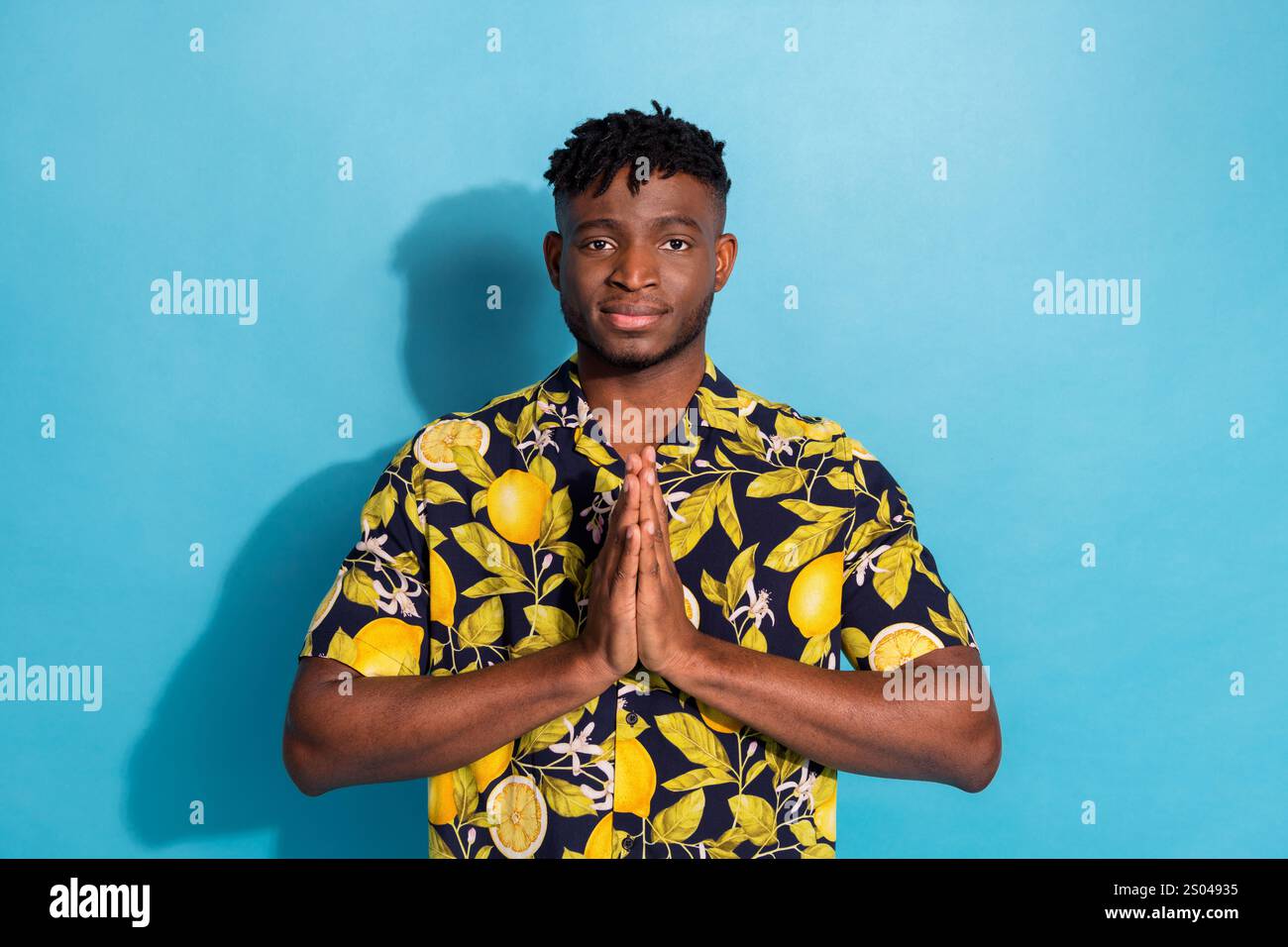 Junger Mann in einem Hemd mit Zitronenmuster steht vor einem leuchtend blauen Hintergrund, Hände zusammen in einer ruhigen Pose und verkörpert Sommermode Stockfoto