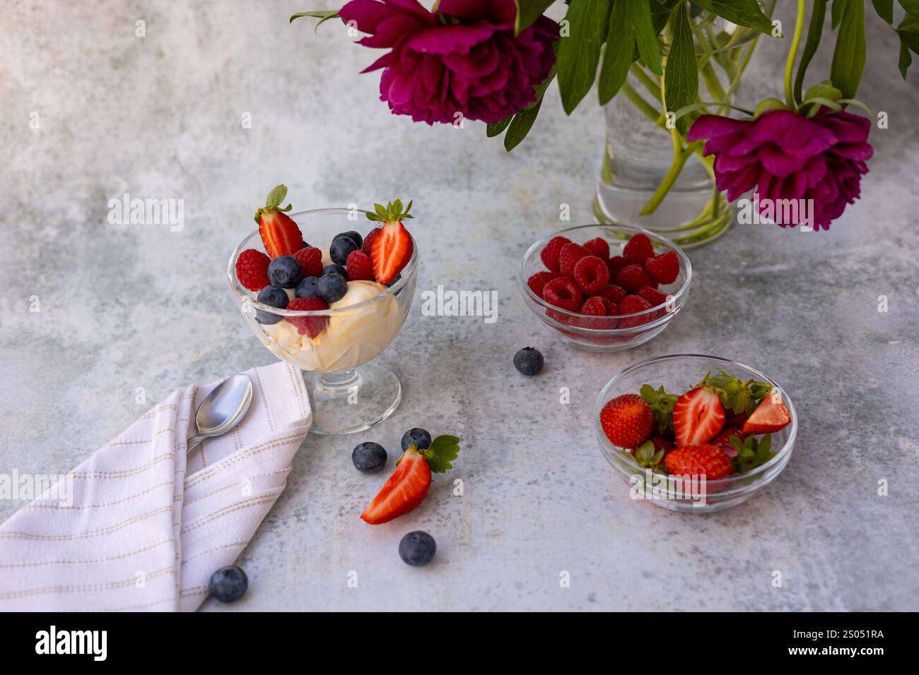 Frisches Sommerdessert mit cremigem Vanilleeis garniert mit Reifen Beeren in einer Glasschale, begleitet von rosa Pfingstrosen auf einer strukturierten Oberfläche Stockfoto