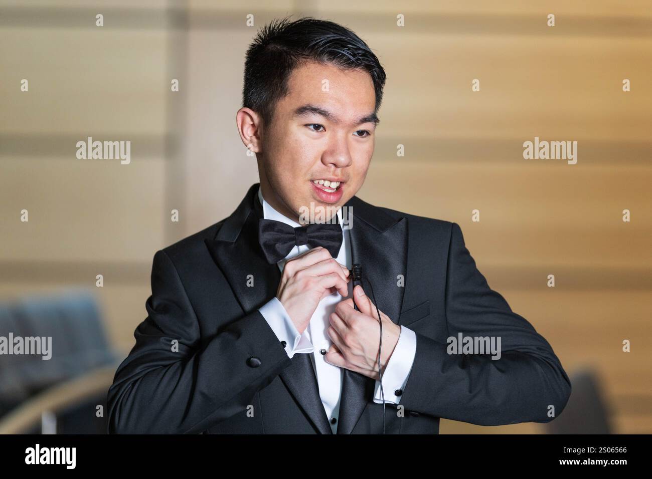 Ein junger Mann in einem Smoking, der während einer formellen Veranstaltung an einem Lavaliermikrofon in einem Auditorium hängt, für ein Interview, in Vorbereitung auf eine Rede Stockfoto