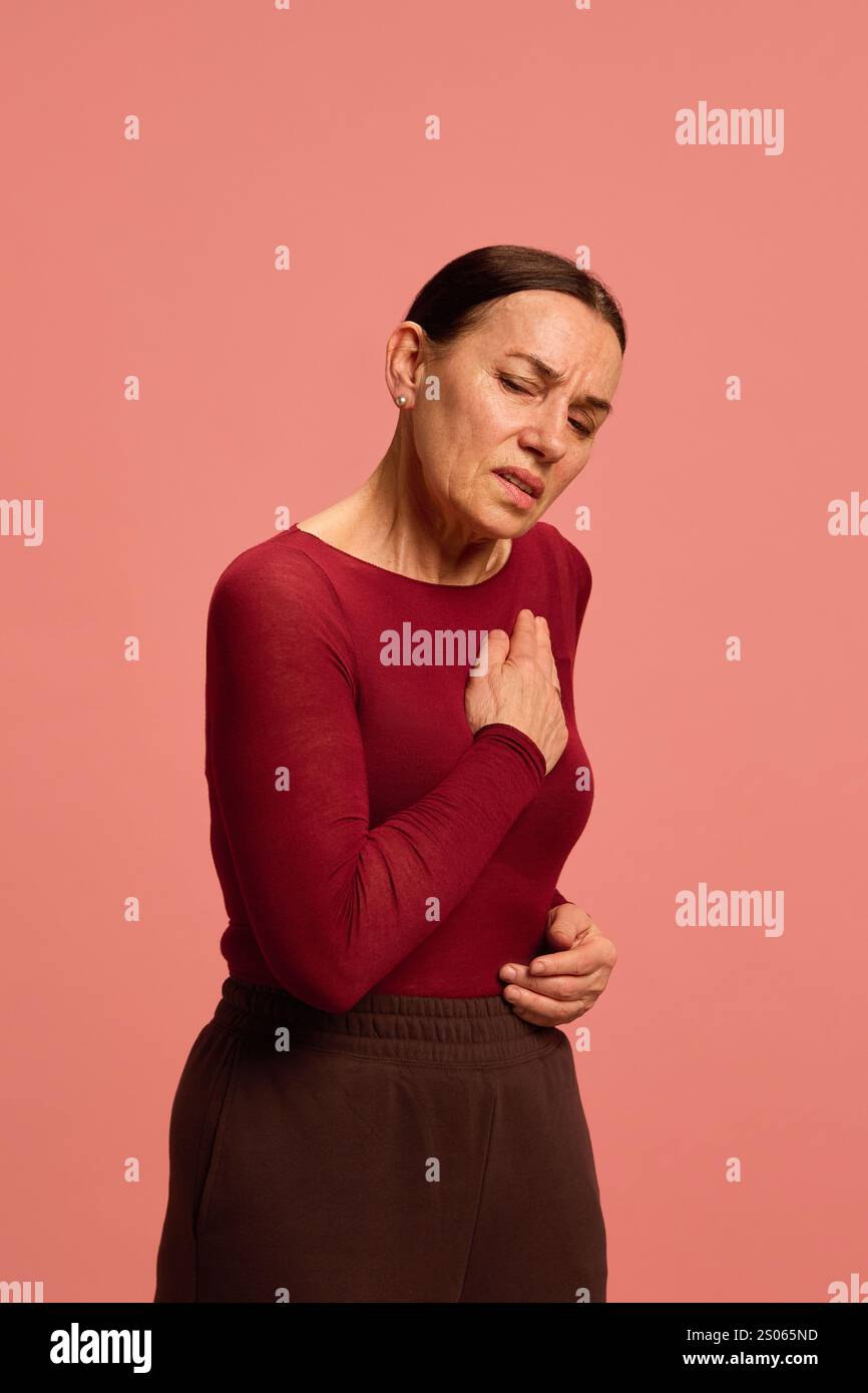 Ältere Frau, Rentnerin umklammert ihre Brust unangenehm, schildert Herzschmerzen oder Kurzatmigkeit vor rosafarbenem Studiohintergrund. Stockfoto