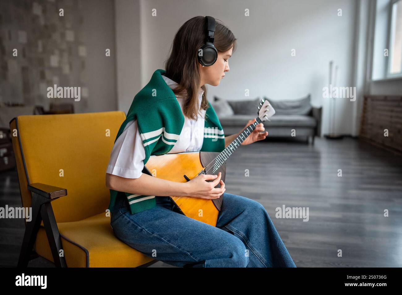 Eine fokussierte Frau spielt Balalaika, fühlt Musik zu Hause über Kopfhörer, übt das Zupfen von Saiten Stockfoto