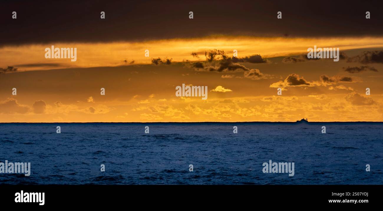 Eine ruhige Meereslandschaft bei Sonnenuntergang mit einem ruhigen Ozean unter einem dramatischen Himmel voller Wolken. Der Horizont leuchtet in warmen goldenen Tönen, während ein kleiner bo Stockfoto