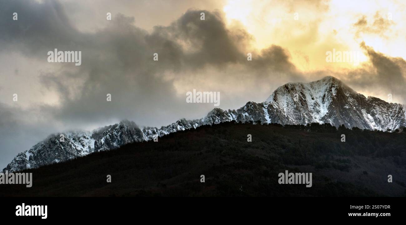Eine dramatische Berglandschaft mit schneebedeckten Gipfeln unter bewölktem Himmel mit einem goldenen Farbton der untergehenden Sonne. Stockfoto