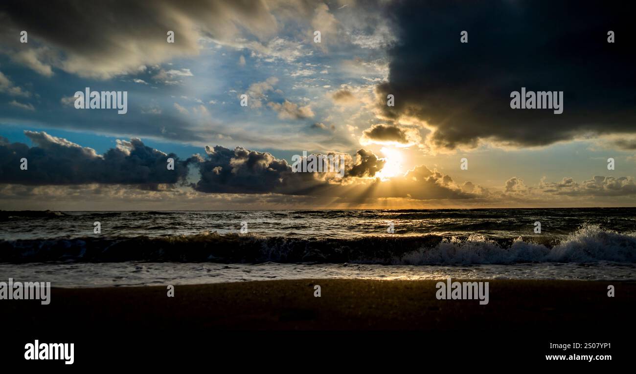 Eine ruhige Strandszene bei Sonnenuntergang, mit Wellen, die sanft am Ufer plätschern. Der Himmel ist voller dramatischer Wolken, und Sonnenstrahlen brechen durch, il Stockfoto