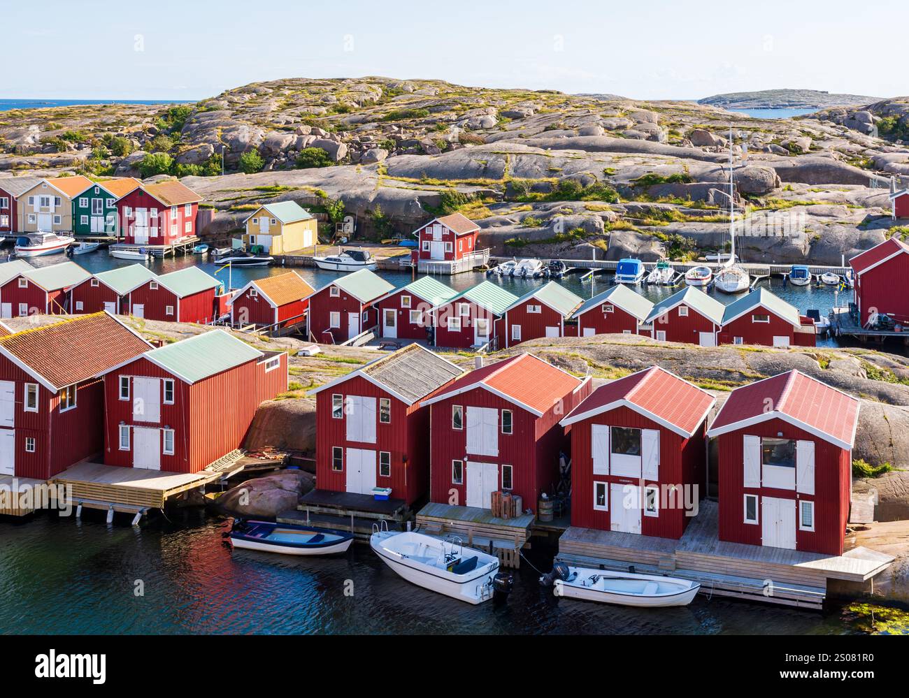 Reihen von rot bemalten Holzhäusern entlang des Meereskanals in Smögen, Schweden, mit der Granitküste in der Ferne an einem sonnigen Sommertag. Stockfoto