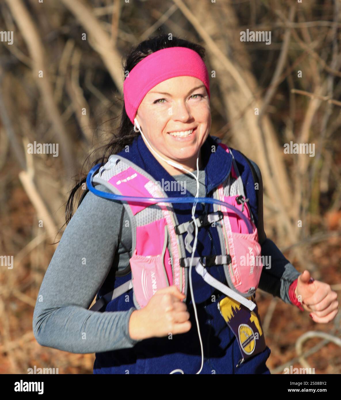 Smithtown, New York, USA - 14. Dezember 2024: Jogger läuft auf einem bewaldeten Pfad, trägt an einem sonnigen Tag ein rosafarbenes Stirnband und eine Feuchtigkeitsweste. Stockfoto