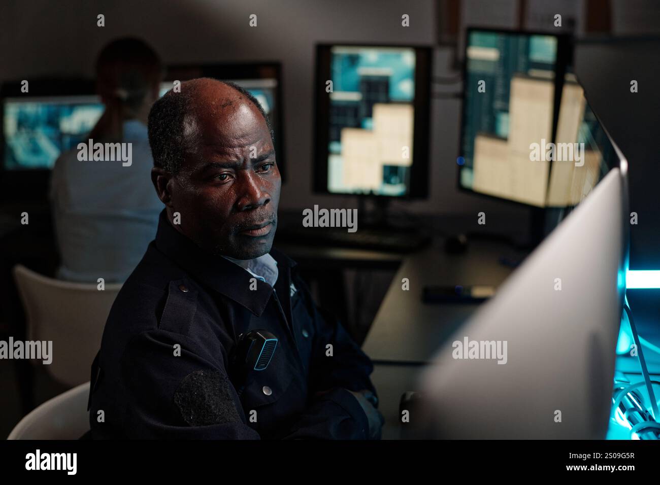 Seriöser, ausgereifter Sicherheitsbeamter sitzt am Arbeitsplatz im Beobachtungsraum und blickt auf einen der Desktop-Computer-Bildschirme Stockfoto
