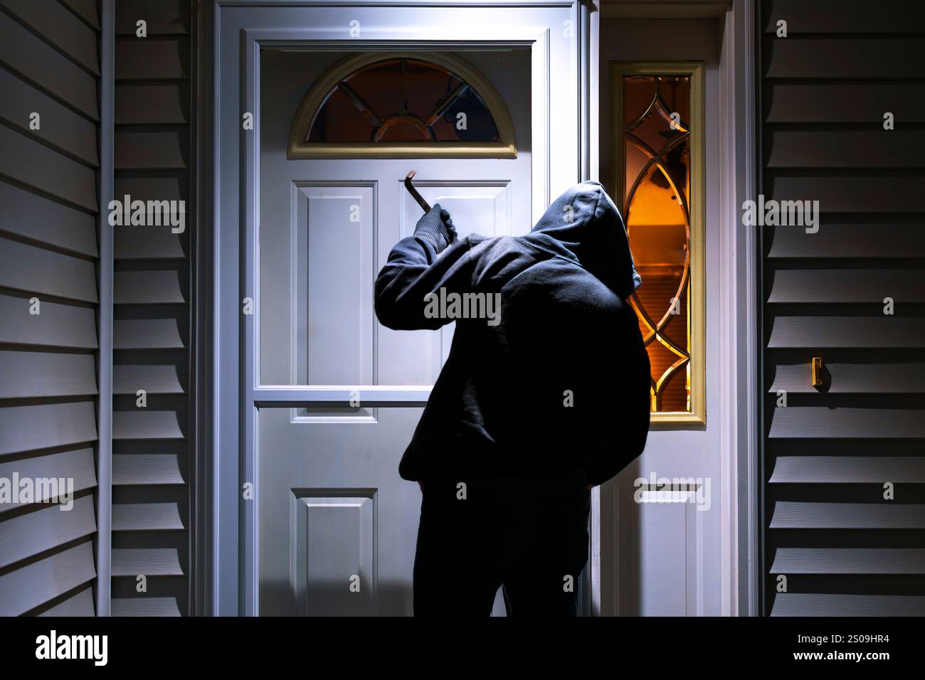Ein Mann in einem Hoodie-Sweatshirt hält eine Brechstange, während er vorgibt, abends in ein Vorstadthaus einzubrechen. Stockfoto
