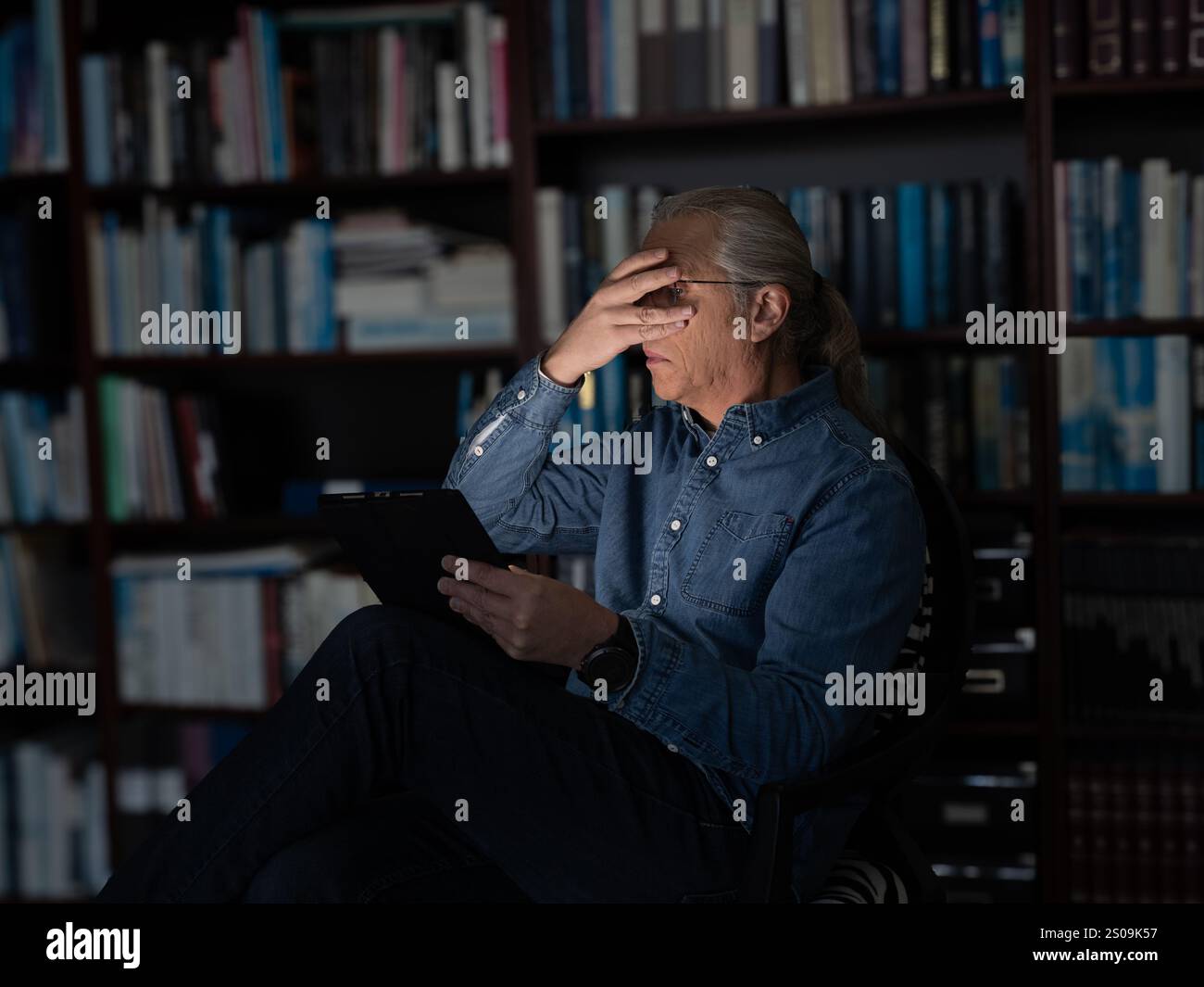 Ein Mann in einem blauen Hemd liest ein unheimliches E-Book, seine Hand erhoeht, um sein beleuchtetes Gesicht abzuschirmen, und ein Bücherregal im Hintergrund verleiht der sce Tiefe Stockfoto