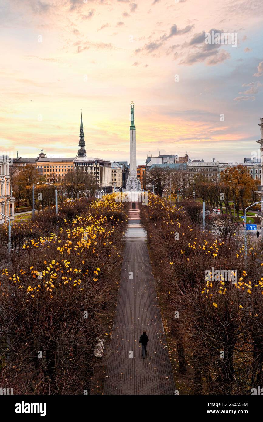 Aus der Vogelperspektive von Riga, Lettland, mit dem Freiheitsdenkmal inmitten des Herbstlaub. Eine Einzelfigur läuft auf einem Weg mit dem Turm der Peterskirche Stockfoto