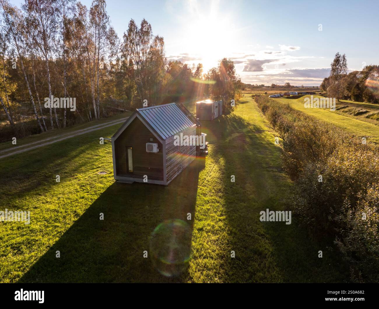 Eine kleine moderne Hütte mit einem Metalldach liegt auf einem grasbewachsenen Bereich. Birkenbäume säumen die linke Seite, mit offenen Feldern, die sich bis zum Horizont unter einem warmen G erstrecken Stockfoto