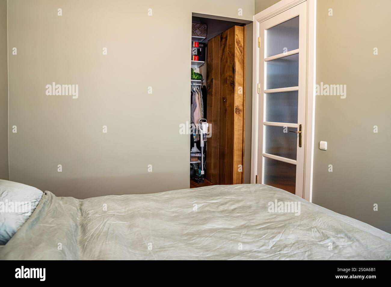 Ein einfaches Schlafzimmer verfügt über ein ordentlich gefertigtes Bett, einen offenen Schrank mit hängenden Kleidern und eine weiße Tür mit mattierten Glaspaneelen, die die Funktionalität betonen. Stockfoto