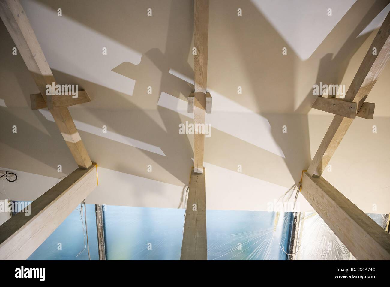 Moderne Innendecke mit Holzbalken, die geometrische Muster bilden. Diffuses Licht und Reflexionen deuten auf große Fenster oder Glasplatten hin. Stockfoto