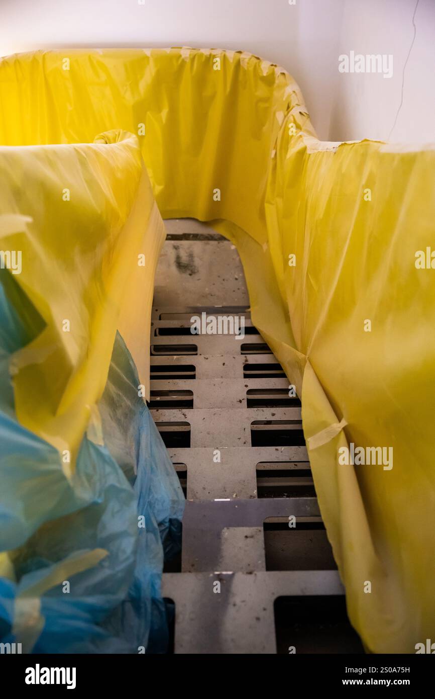 Ein schmaler Innenraum verfügt über einen Metallrostboden und eine leuchtend gelbe und blaue Kunststofffolie, was auf eine Bau- oder Renovierungsanlage hindeutet. Stockfoto