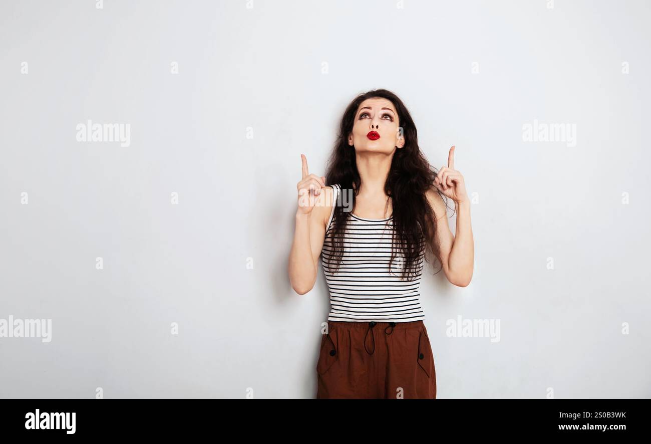 Schöne positive, lustige Make-up-Frau in gestreiftem lässigem T-Shirt, die aufblickt und auftaucht und das Ideenzeichen mit zwei Fingern oben auf leerer Kopie zeigt Stockfoto